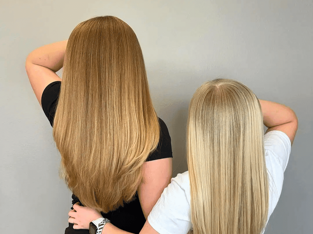 Two people with long, straight hair; one with light brown and one with blonde hair, facing away.