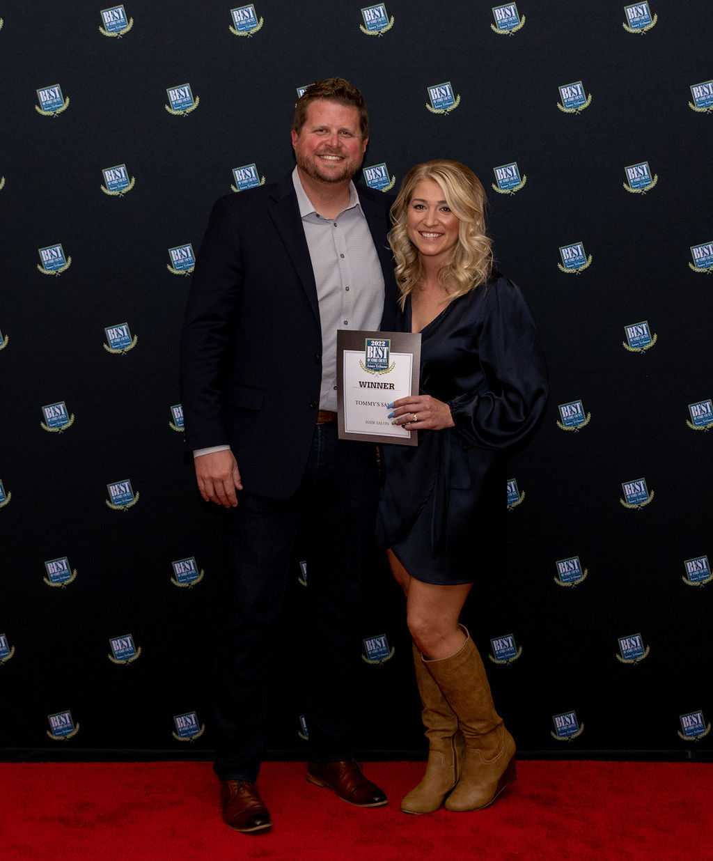 Man and woman celebrating award on a red carpet, holding a "Winner" plaque.