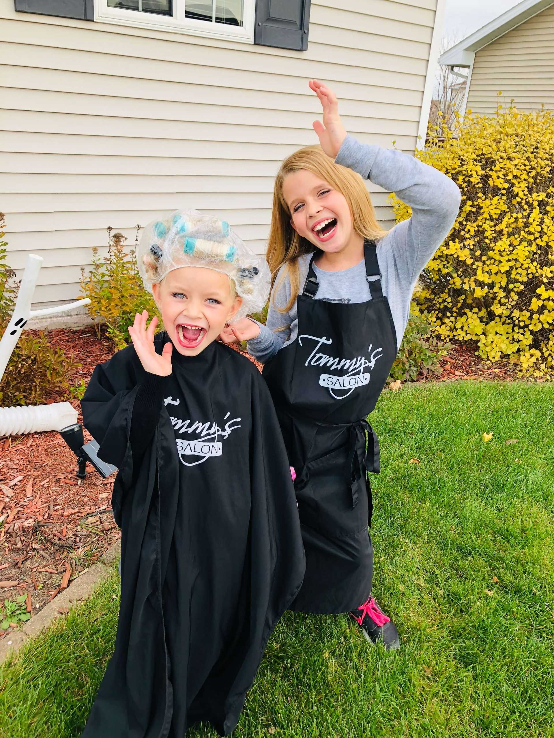 Two girls smiling and posing in aprons outside, one wearing hair curlers.