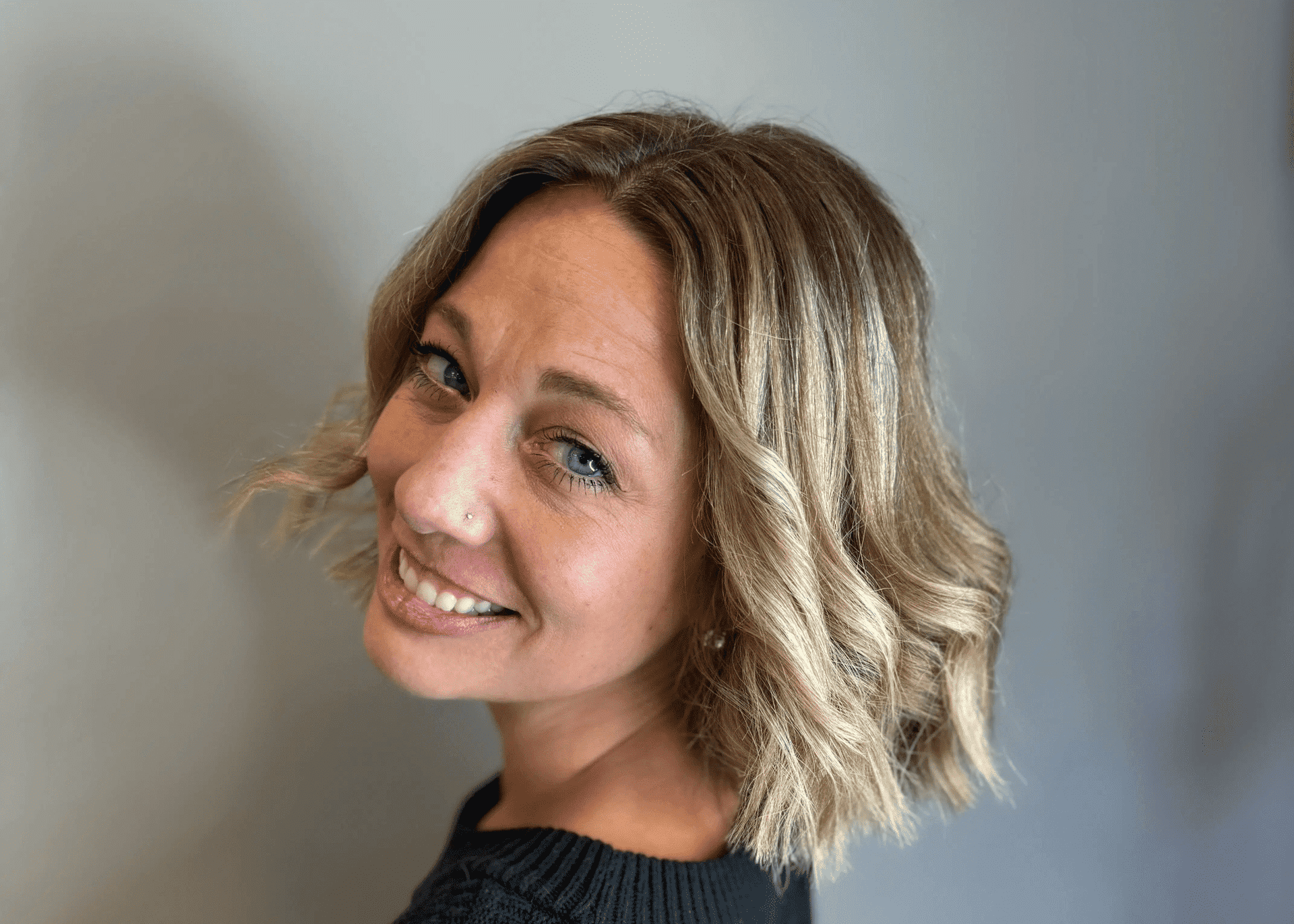 Smiling woman with short blonde hair, wearing a black sweater, looking over her shoulder.